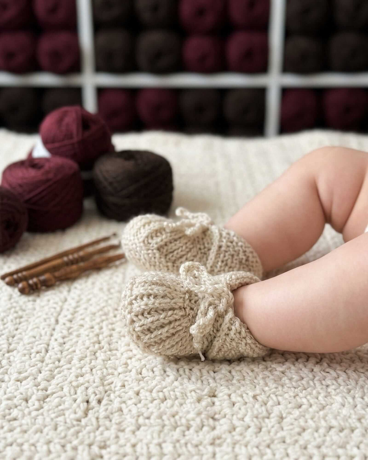 A close-up of a baby's legs wearing beige hand-knit booties with small bows, resting on a cream knit blanket with yarn balls and knitting needles in the background.