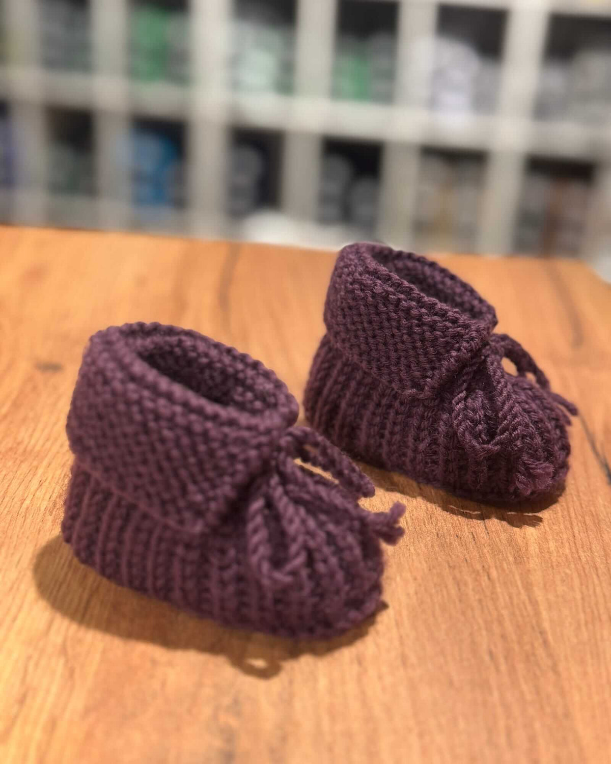 A pair of handmade, knitted baby booties in a deep plum purple, resting on a wooden tabletop with a blurred background.