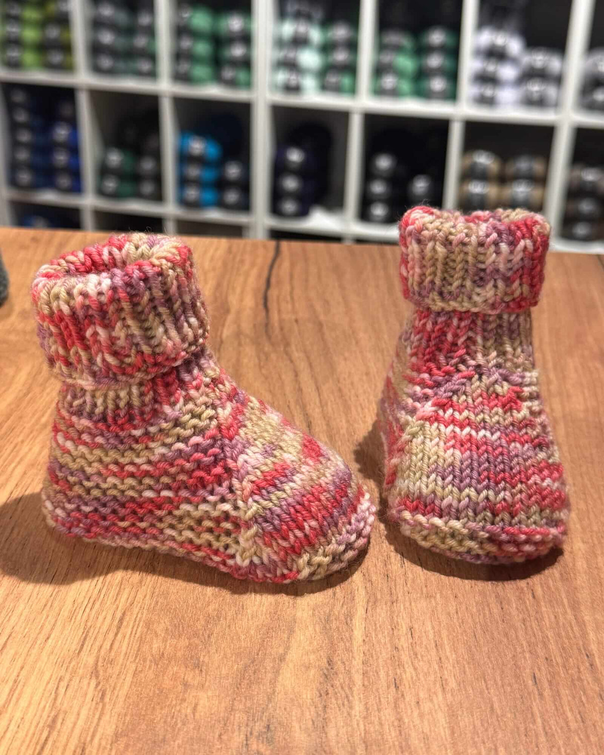 A pair of hand-knit baby booties in a variegated marled yarn of pink, red, beige, and cream, displayed on a wooden tabletop with a blurred background of shelves.