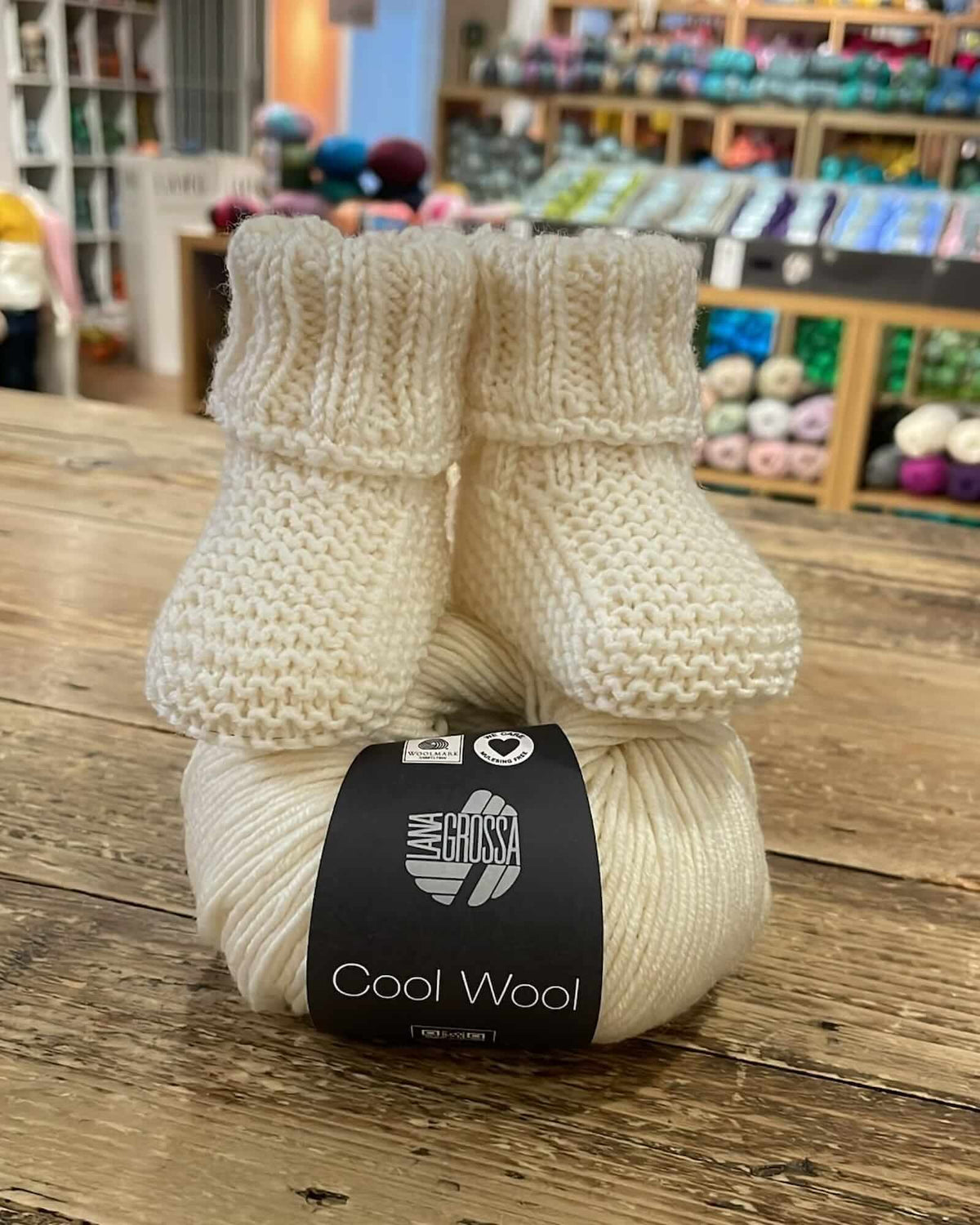 a hand-knitted merino baby booties, placed on a wool yarn —all in a cream color—placed on a wooden table inside a yarn shop