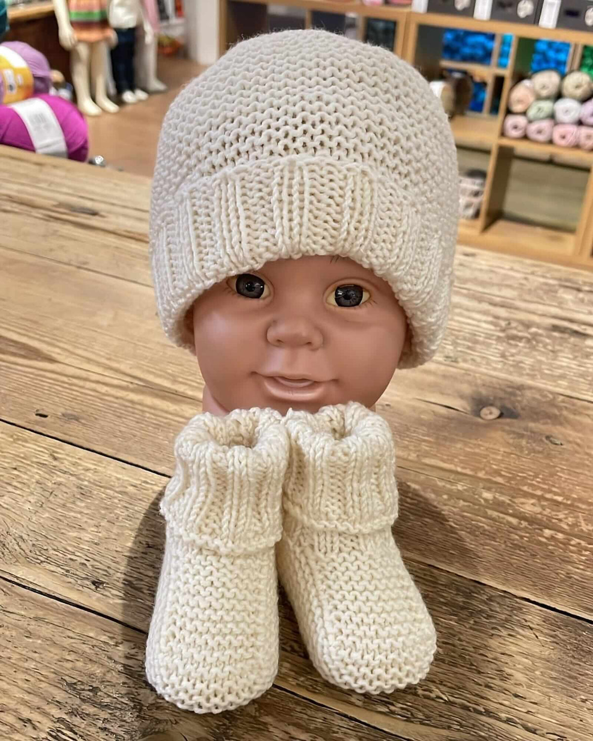 a hand-knitted merino set consisting of booties and a hat, displayed on a mannequin head—all in a cream color—placed on a wooden table inside a yarn shop