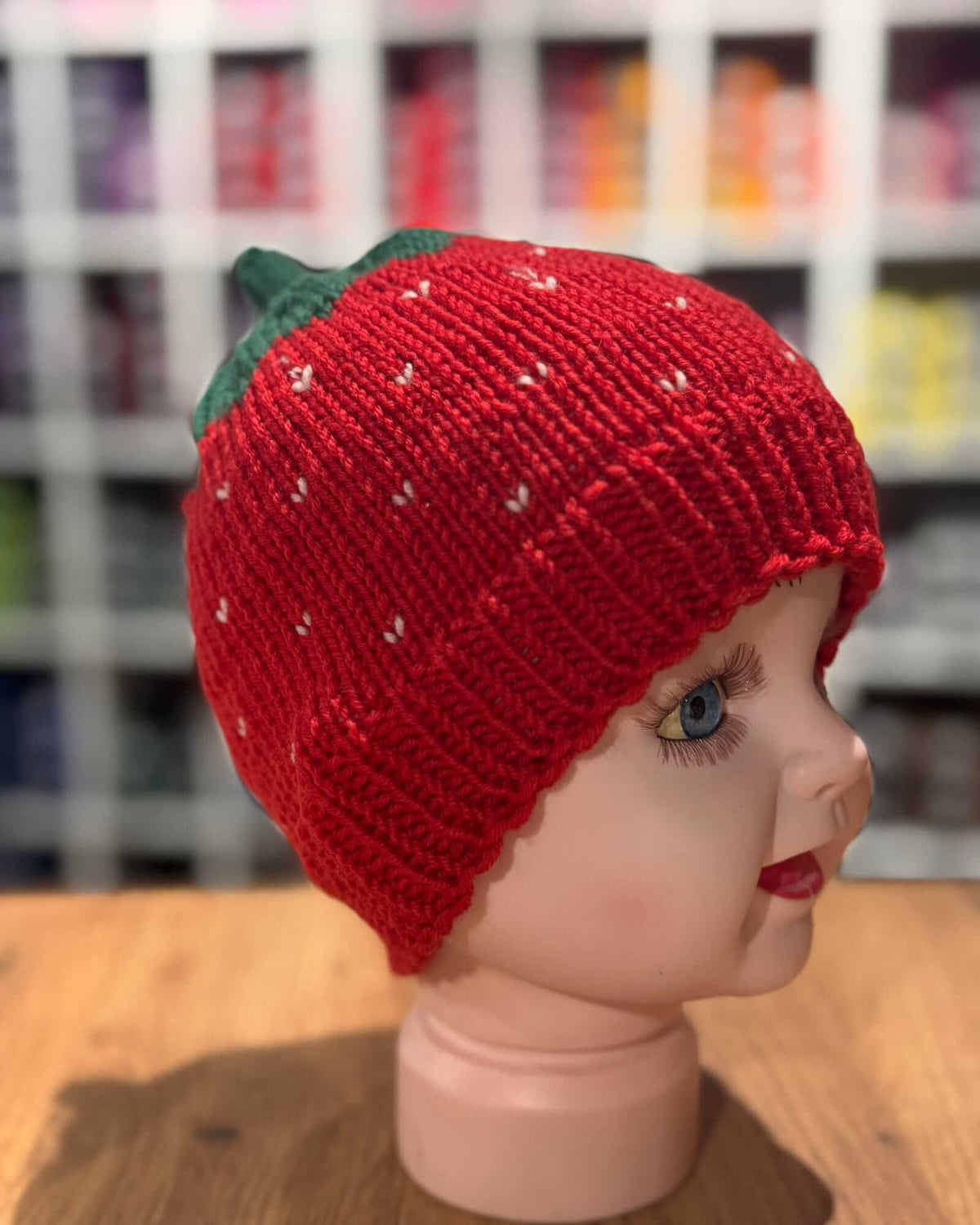 A red knit strawberry hat with a green leaf top, displayed on a mannequin head in a store setting with blurred shelves in the background.