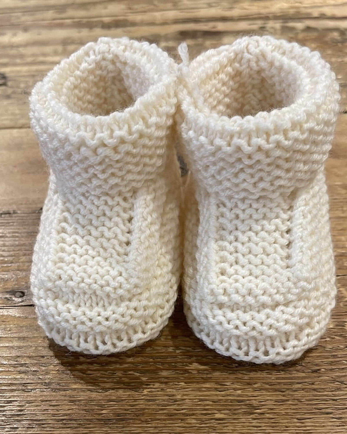 A pair of cream-colored, hand-knit baby booties with a textured knit pattern, displayed on a rustic wooden surface.