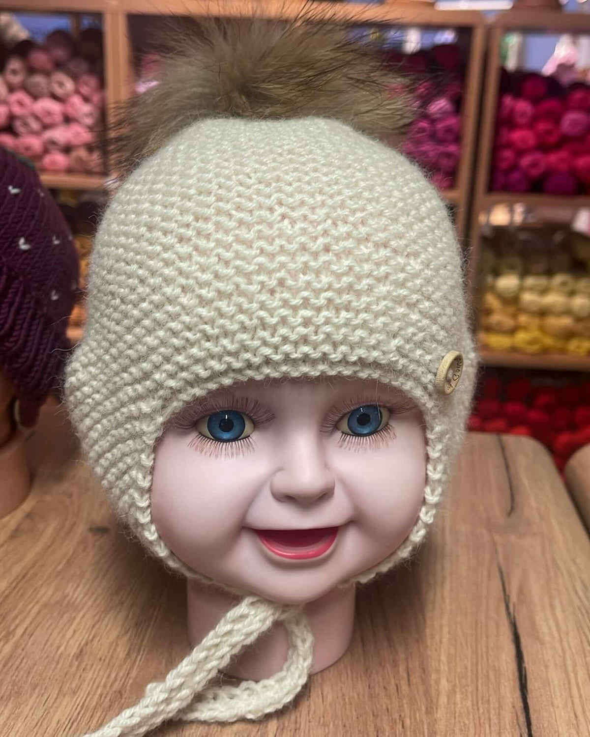 A cream-colored knit baby bonnet with a brown faux fur pom-pom on top, displayed on a doll's head. The bonnet features a small decorative button on the side and a chin strap, set against a background of shelves filled with colorful yarns.