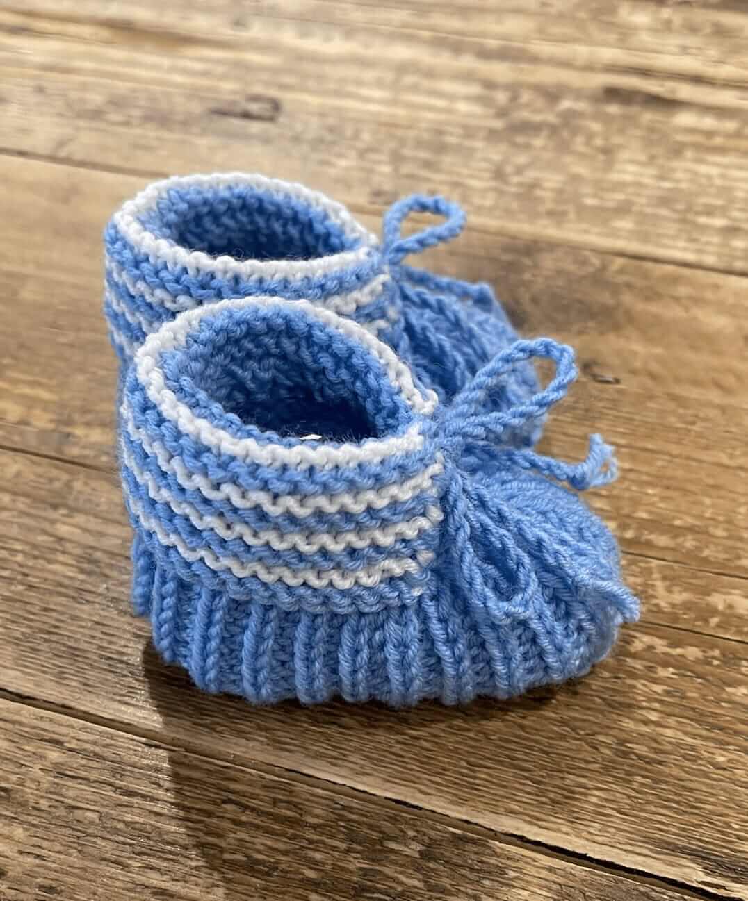 A pair of handmade blue and white striped knit baby booties with ties, displayed on a rustic wooden surface.