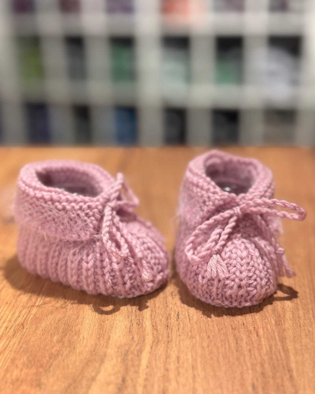 A pair of pink hand-knit baby booties with small bows, resting on a wooden surface with a blurred background.