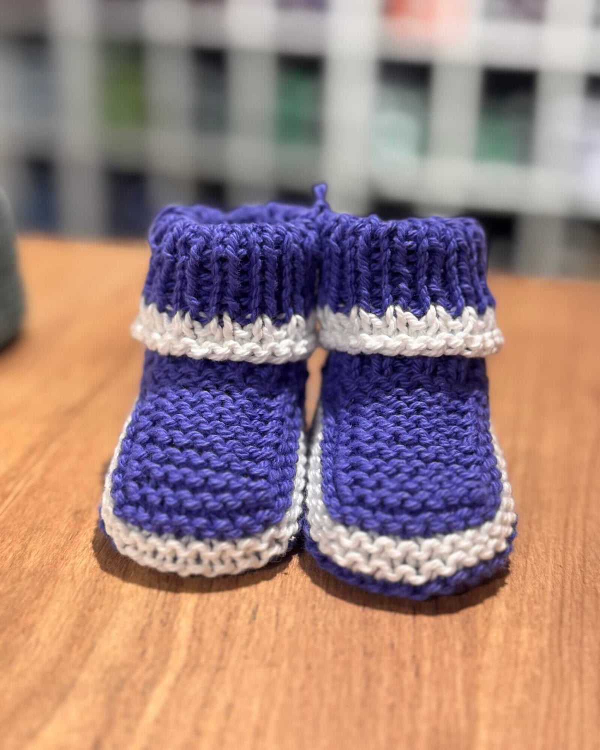 A pair of hand-knit baby booties in purple and white, displayed on a wooden tabletop with a blurred background.