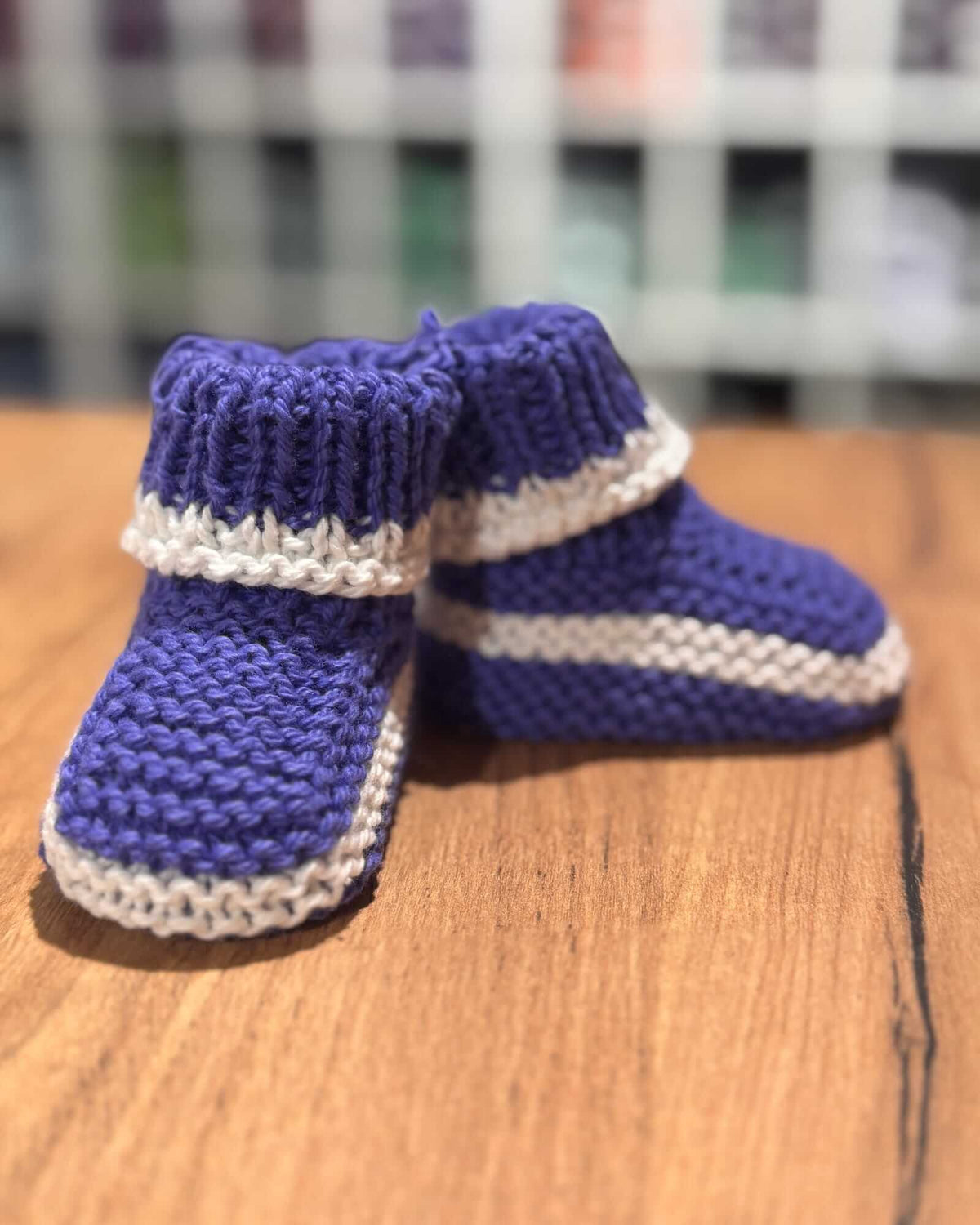 Hand-knitted baby booties in purple and cream, displayed on a wooden surface with a blurred background.