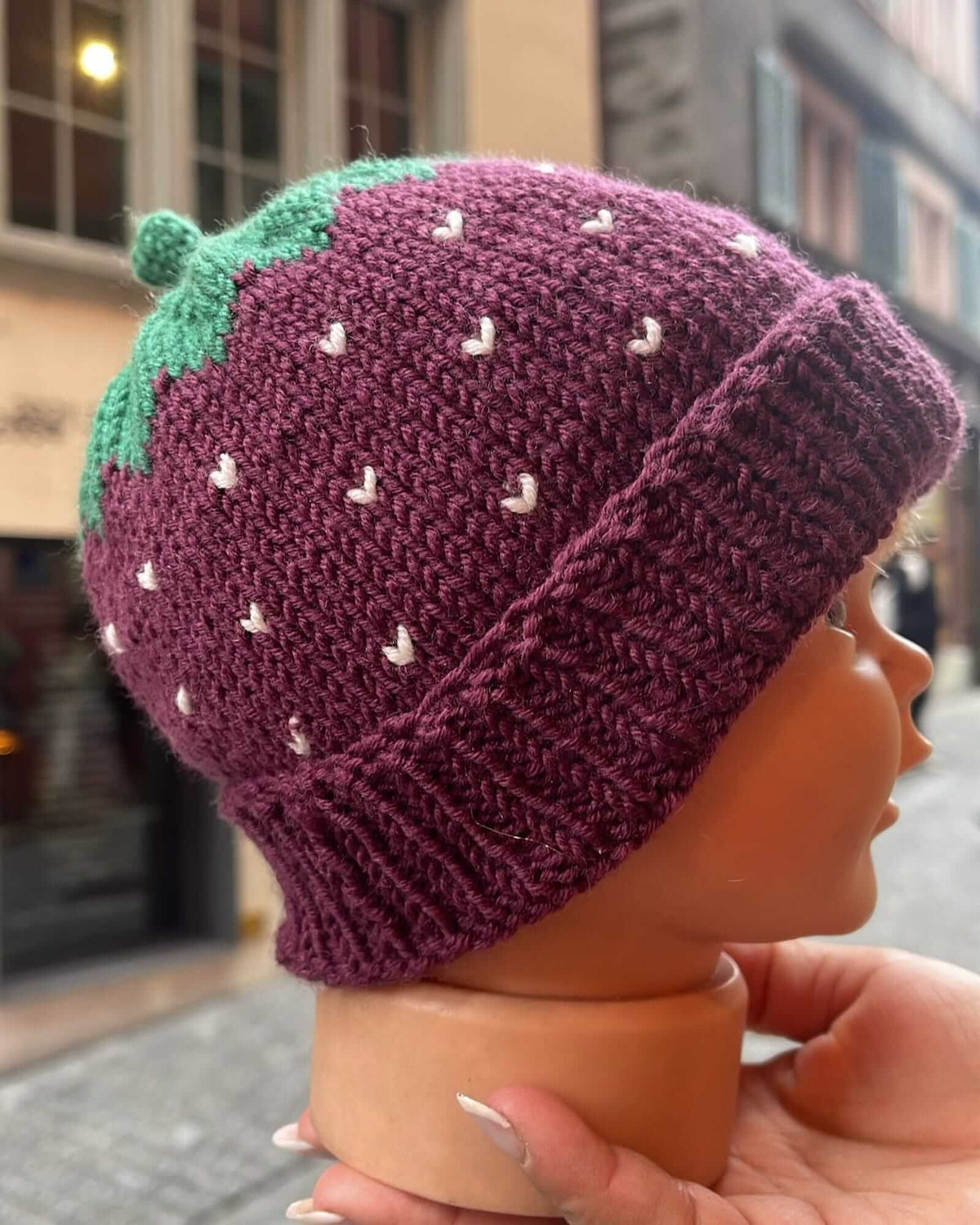 A hand-knit strawberry hat in deep purple with green leafy top and small white seed details, displayed on a mannequin head held by a hand against an outdoor street background.