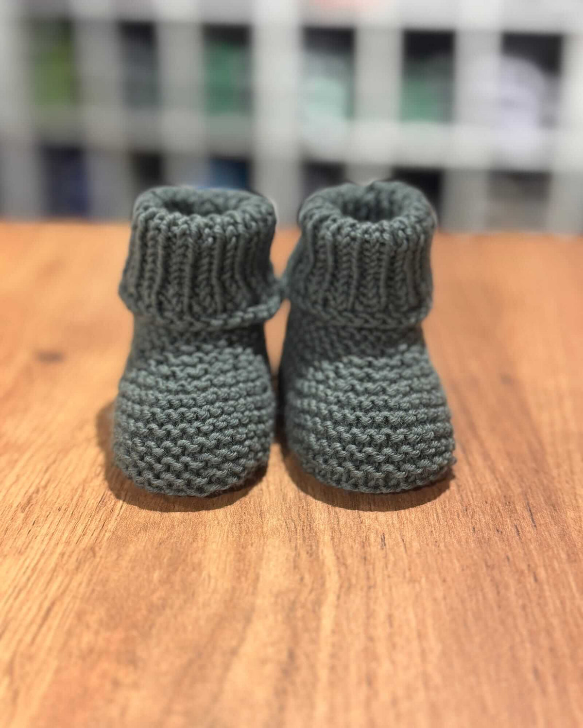 A pair of handmade, ribbed-knit baby booties in a muted sage green, displayed on a wooden tabletop with a blurred background.