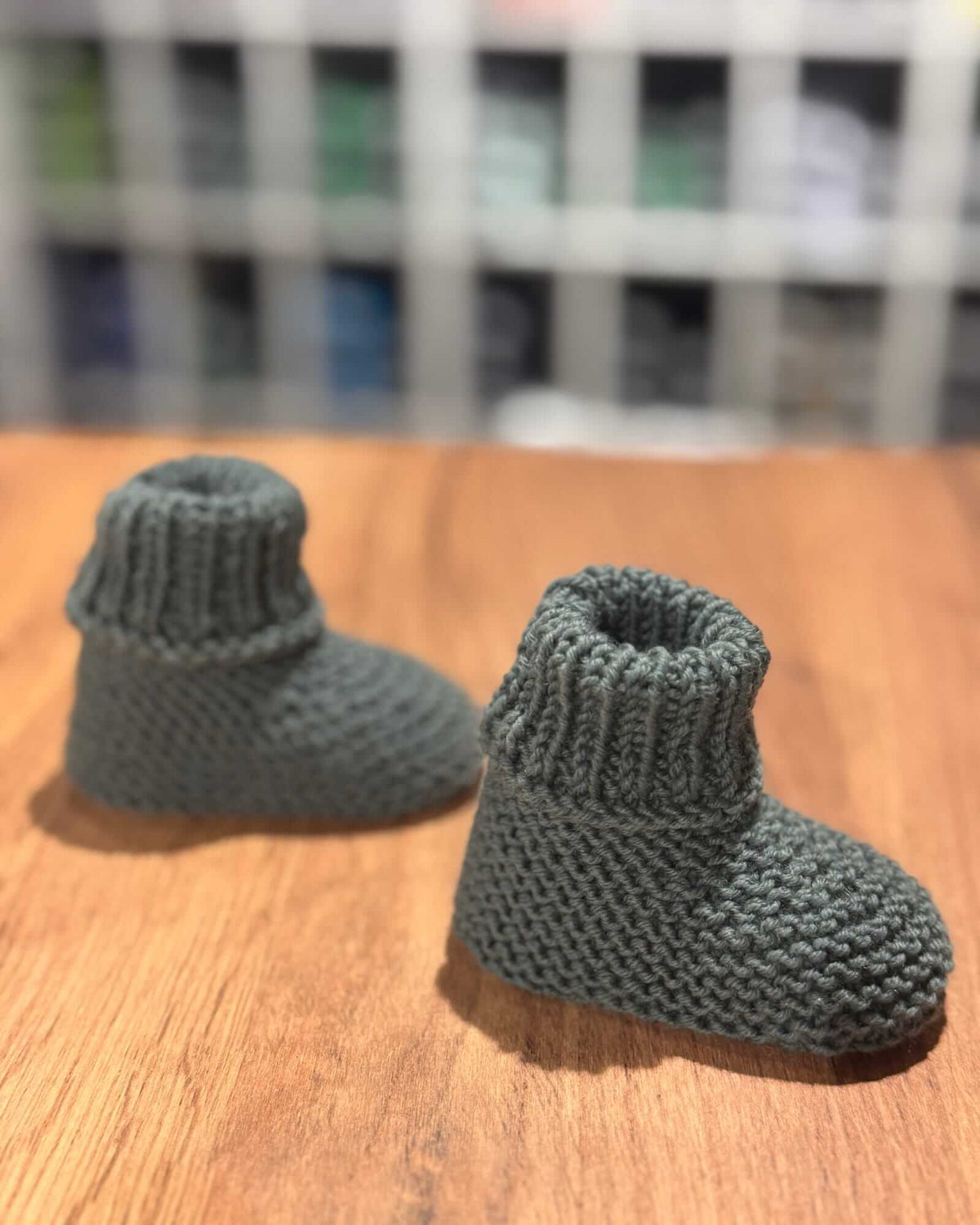 A pair of handmade, dark gray knit baby booties resting on a wooden tabletop with a blurred shelf background.