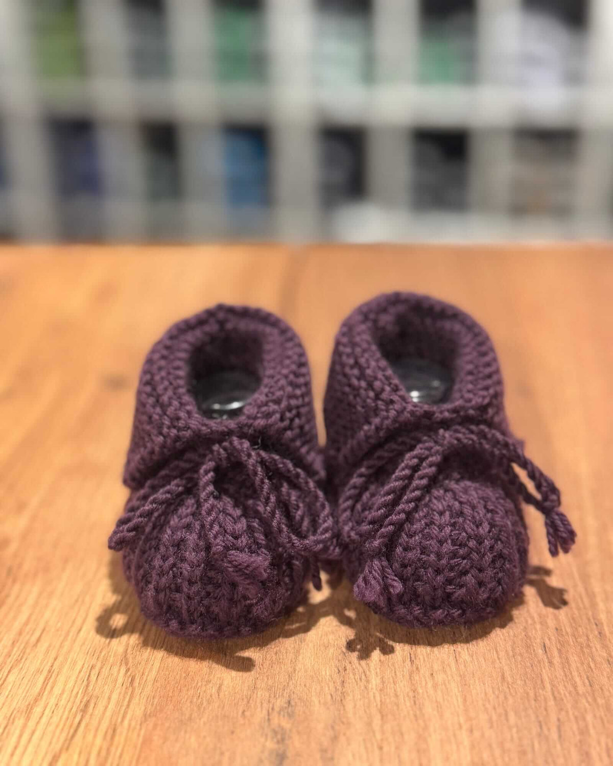 A pair of handmade, dark purple chunky-knit baby booties with matching bow details, resting on a wooden surface with a blurred background.