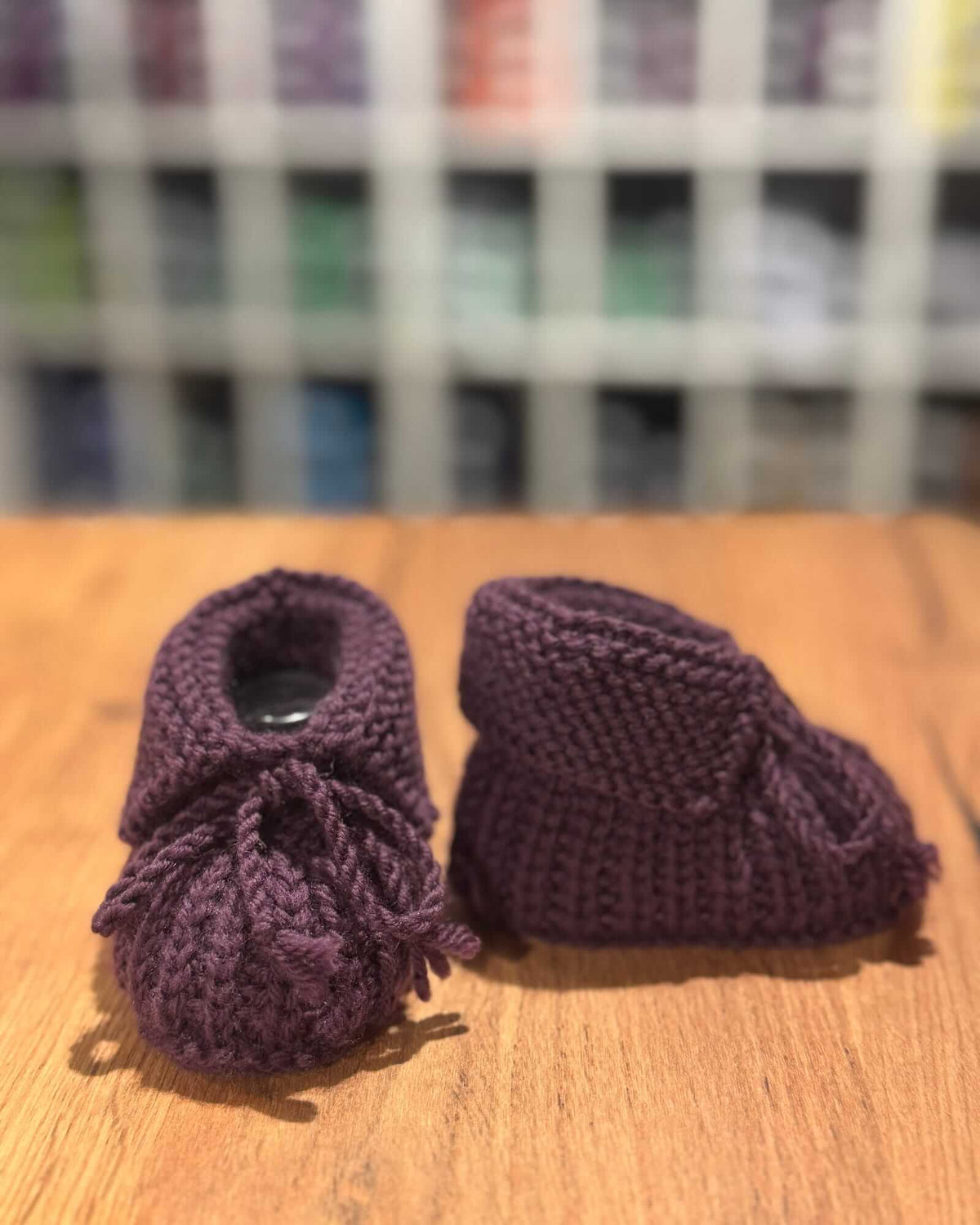 A pair of handmade, hand-knit baby booties in a deep plum purple, displayed on a wooden surface with a blurred background.