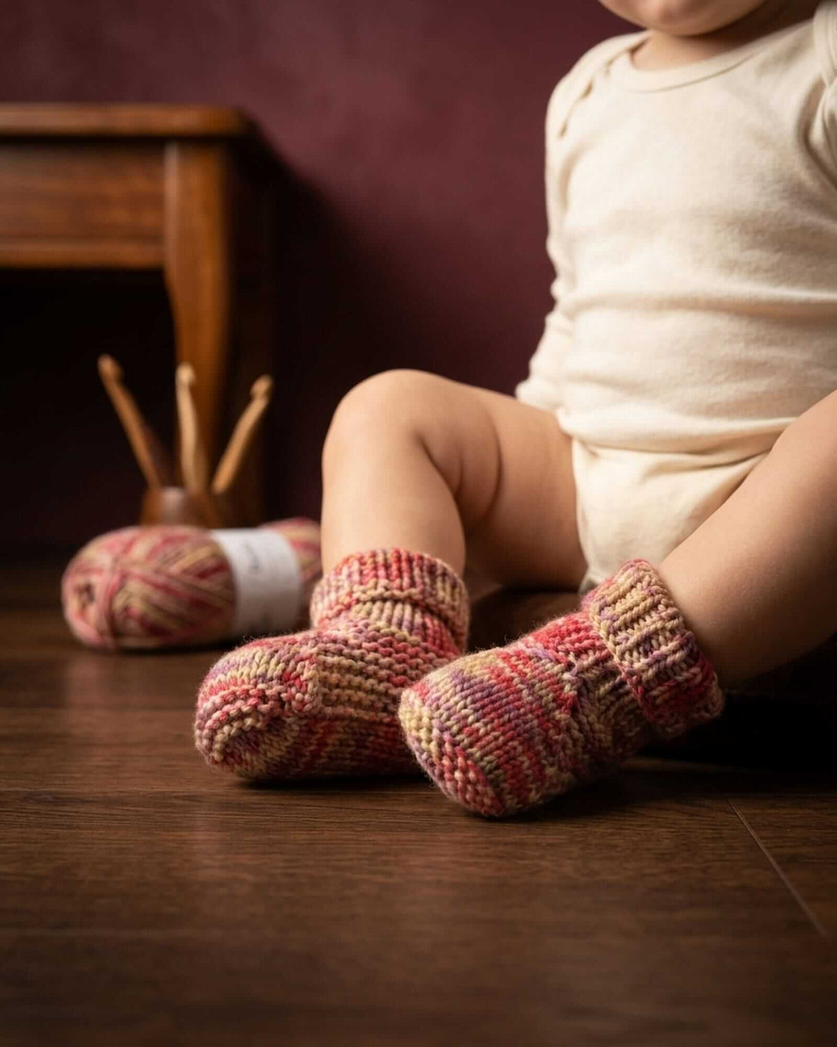 A close-up lifestyle shot of a baby sitting on a wooden floor, wearing hand-knit baby booties in a variegated red, pink, and beige pattern. A ball of matching yarn and knitting needles are visible in the background against a dark burgundy wall.
