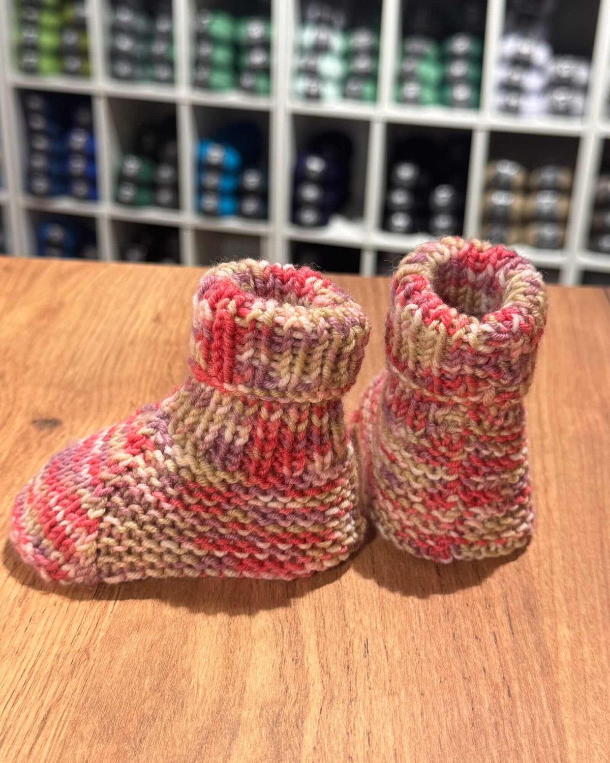 A pair of hand-knit baby booties in a variegated yarn blend of pink, red, beige, and purple, resting on a wooden tabletop with a blurred store shelf in the background.