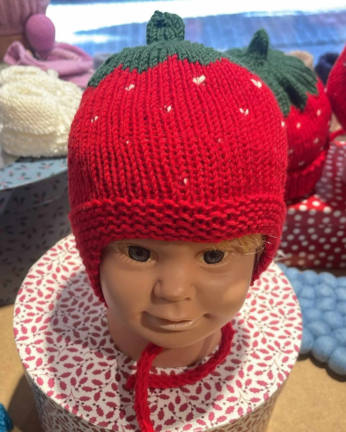 a red strawberry-shaped knit hat with green leaves and long red ear loops placed on a manequinn head