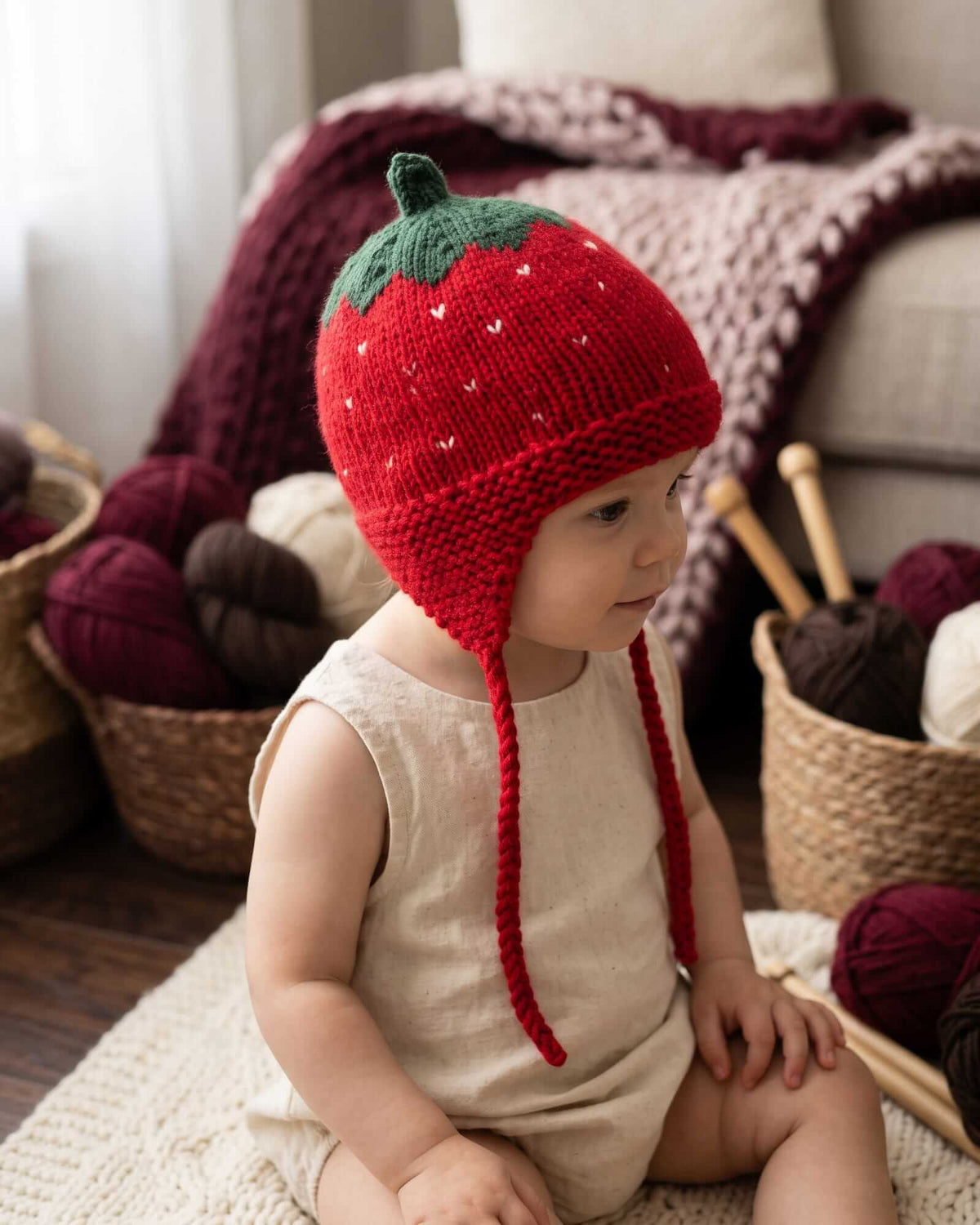 A baby wearing a red strawberry-shaped knit hat with green leaves and long red ear loops, sitting indoors on a rug with baskets of yarn and a blanket in the background.