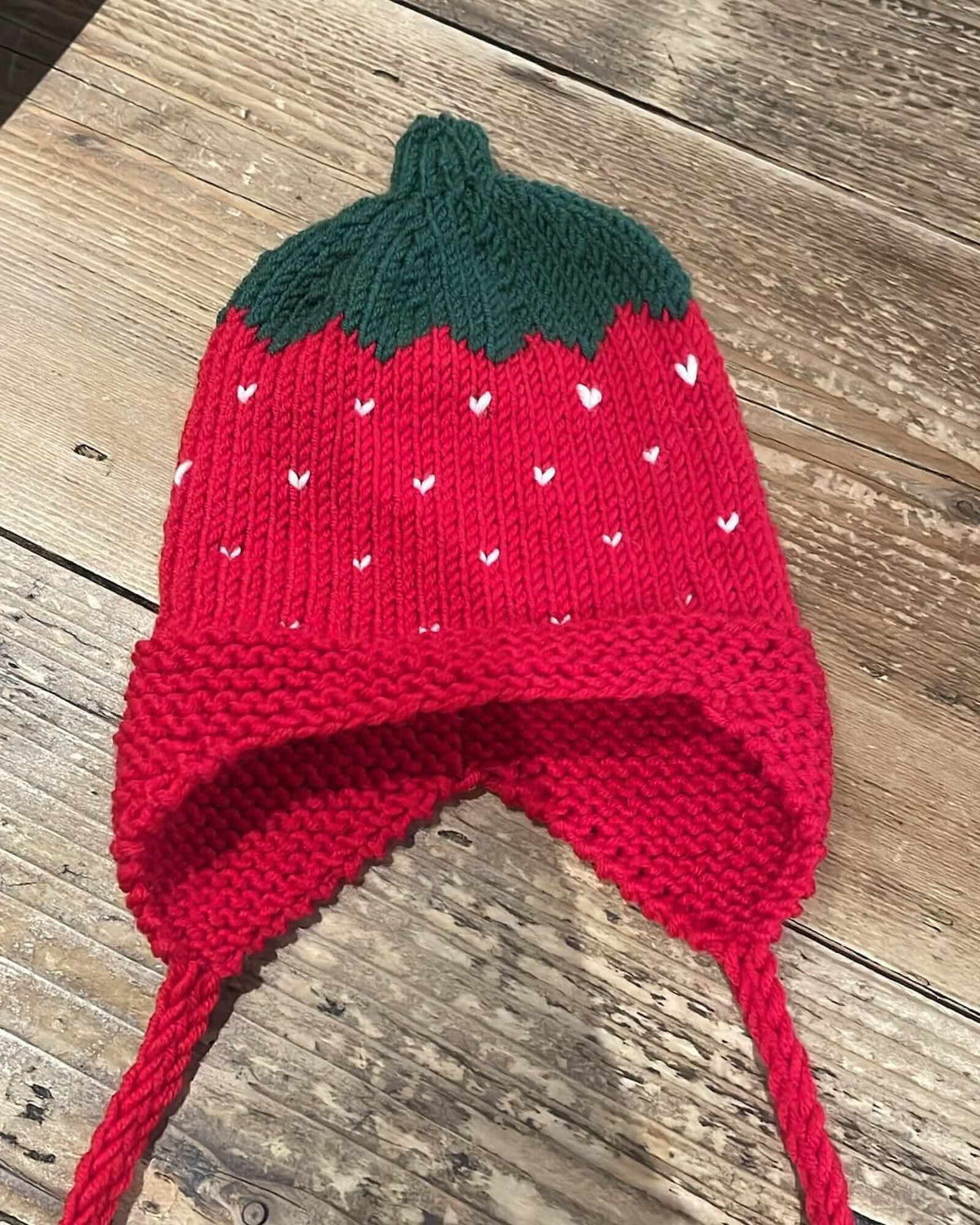 a red strawberry-shaped knit hat with green leaves and long red ear loops placed on a wooden table