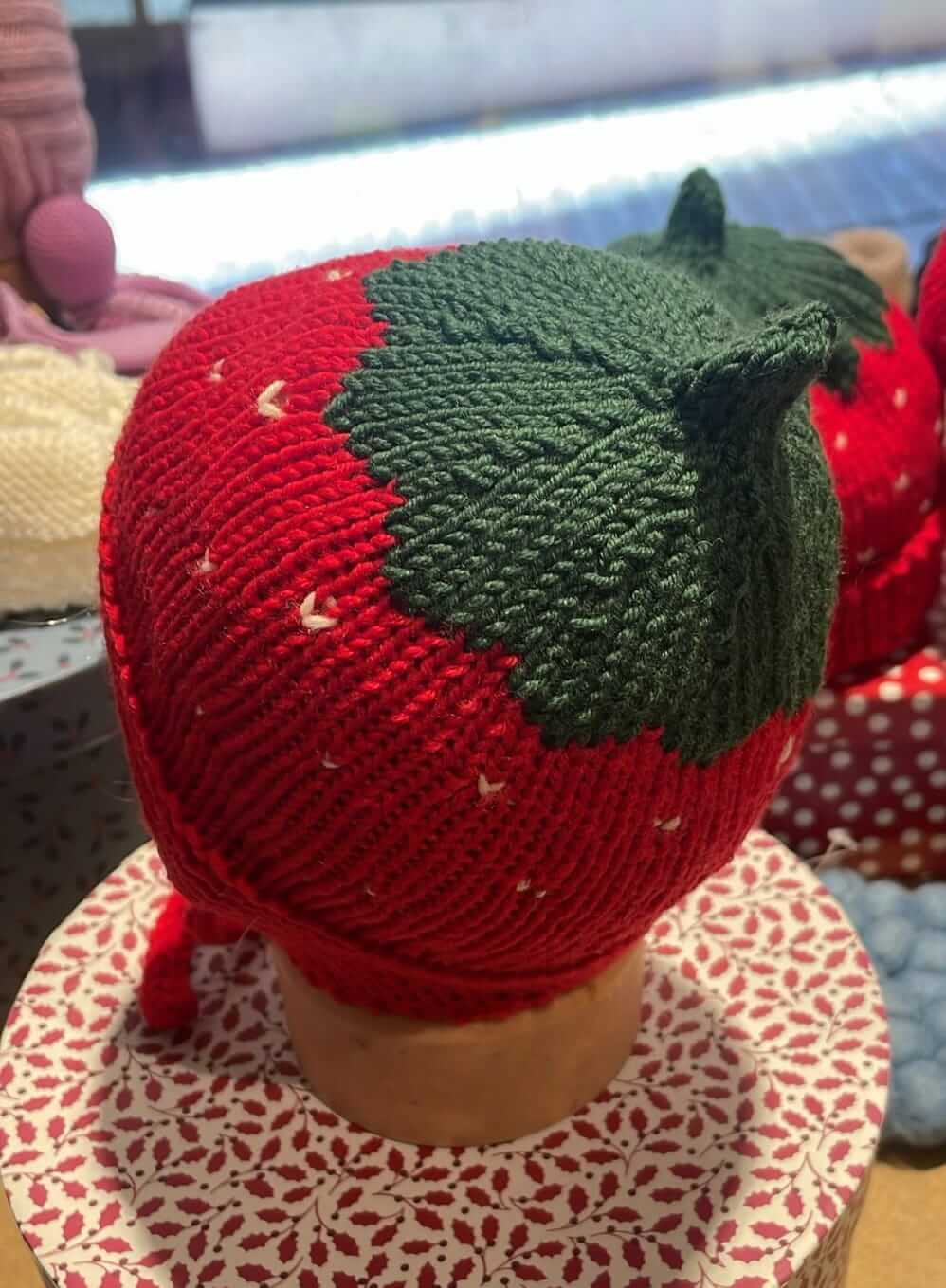 a red strawberry-shaped knit hat with green leaves and long red ear loops on a manequinn-head placed on a gift box