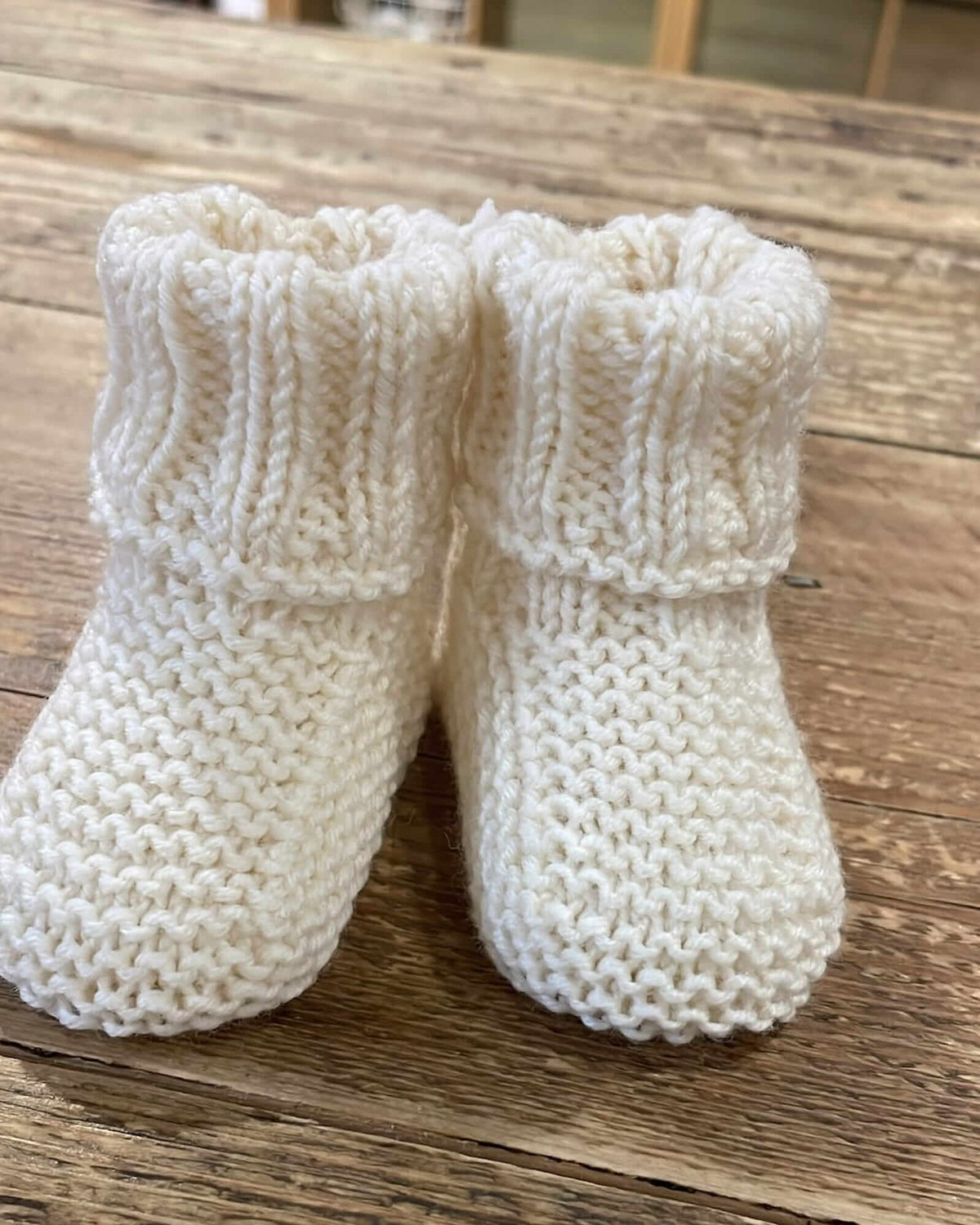 A pair of cream-colored, hand-knit baby booties with a ribbed cuff, resting on a rustic wooden surface.