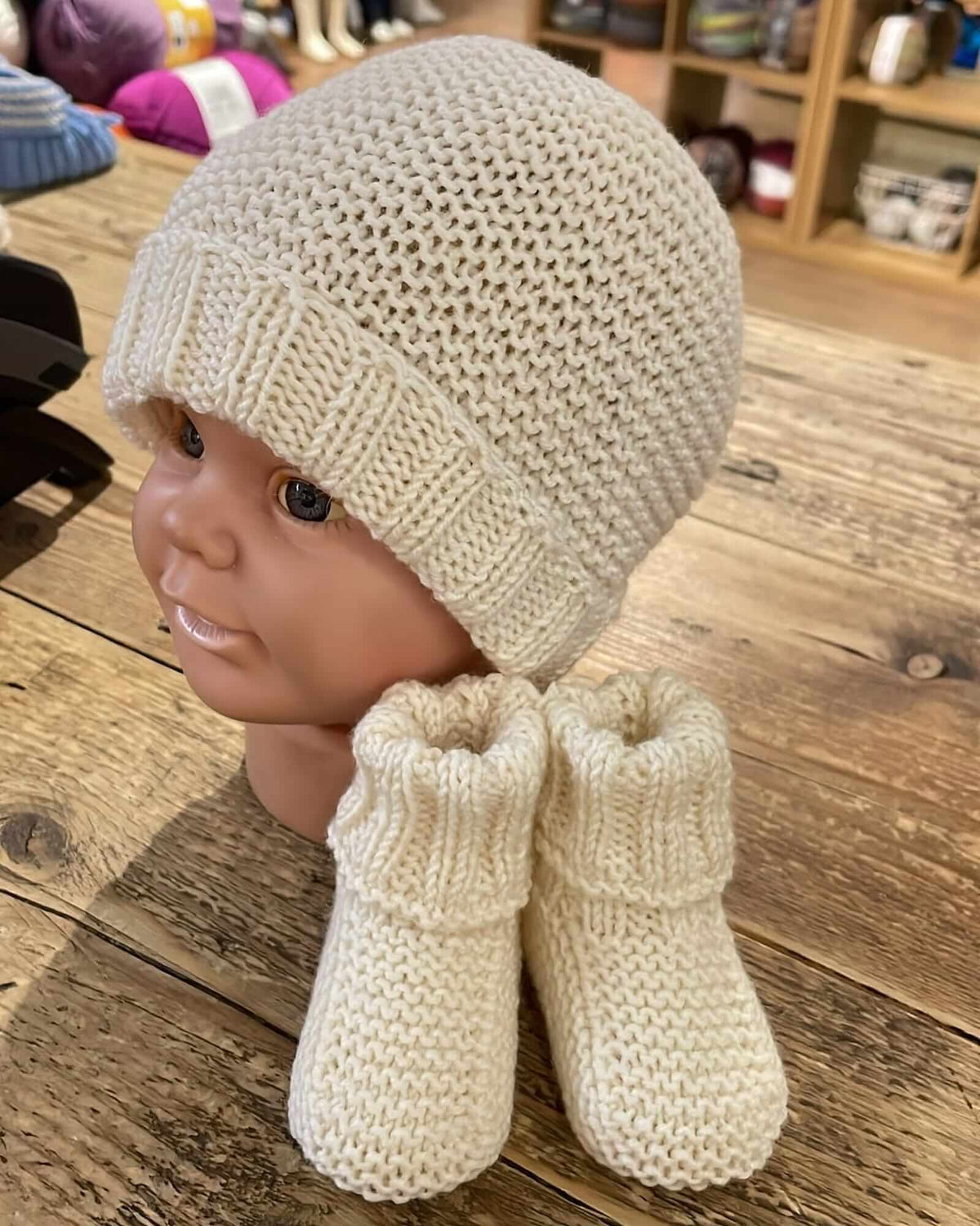 a hand-knitted merino set consisting of booties and a hat, displayed on a mannequin head—all in a cream color—placed on a wooden table inside a yarn shop