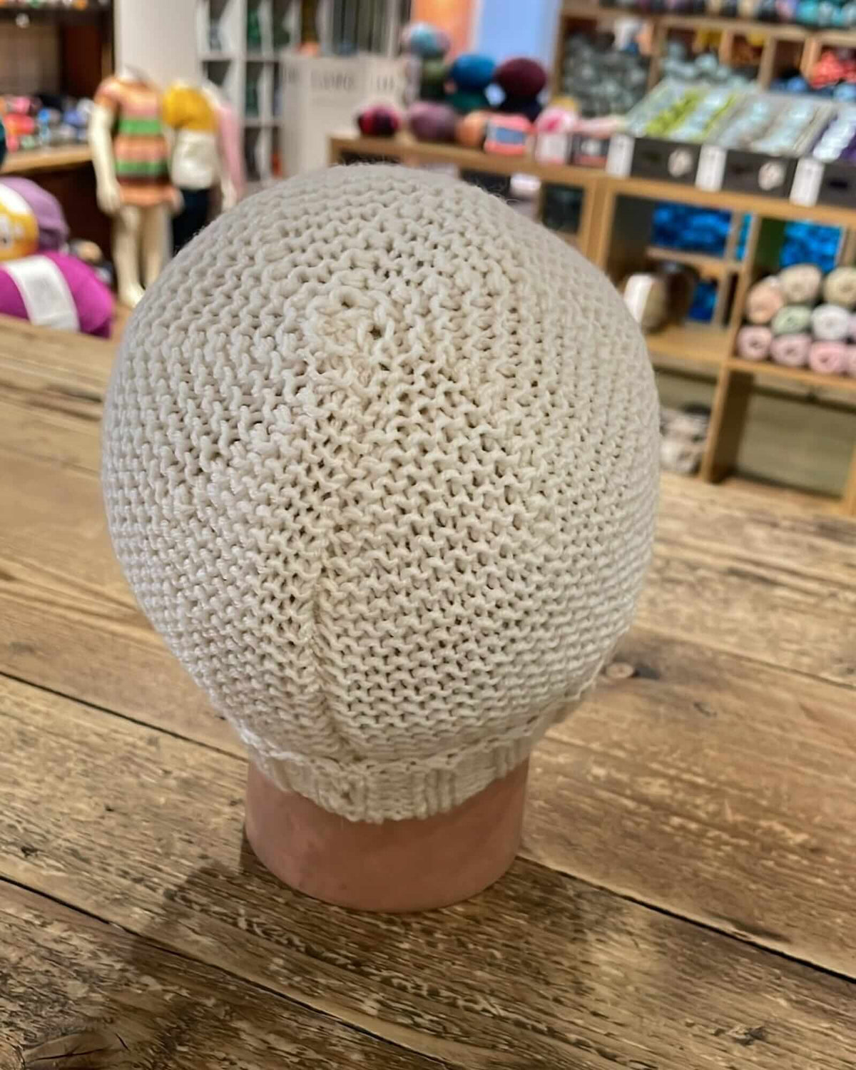 a hand-knitted beige beanie hat shown from behind, placed on a manequinn head, sitting on a wooden table inside a yarn store