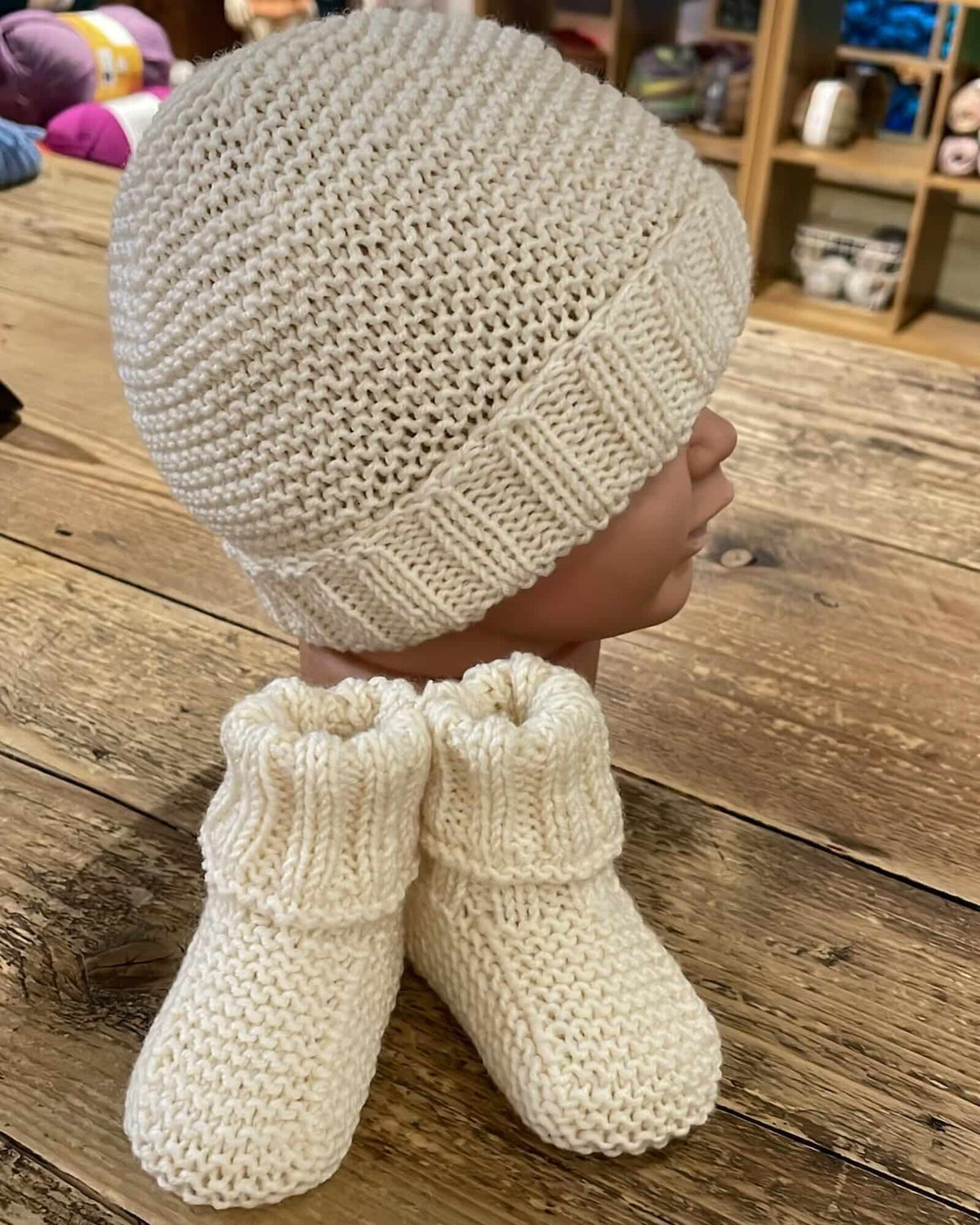 a hand-knitted merino set consisting of booties and a hat, displayed on a mannequin head—all in a cream color—placed on a wooden table inside a yarn shop