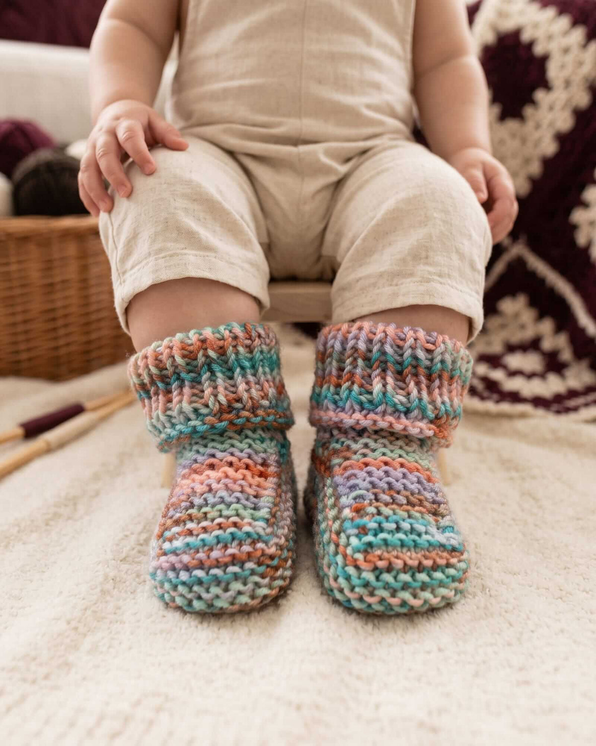A close-up of a baby wearing hand-knitted multicolor booties, paired with a beige linen romper, sitting on a soft cream carpet.