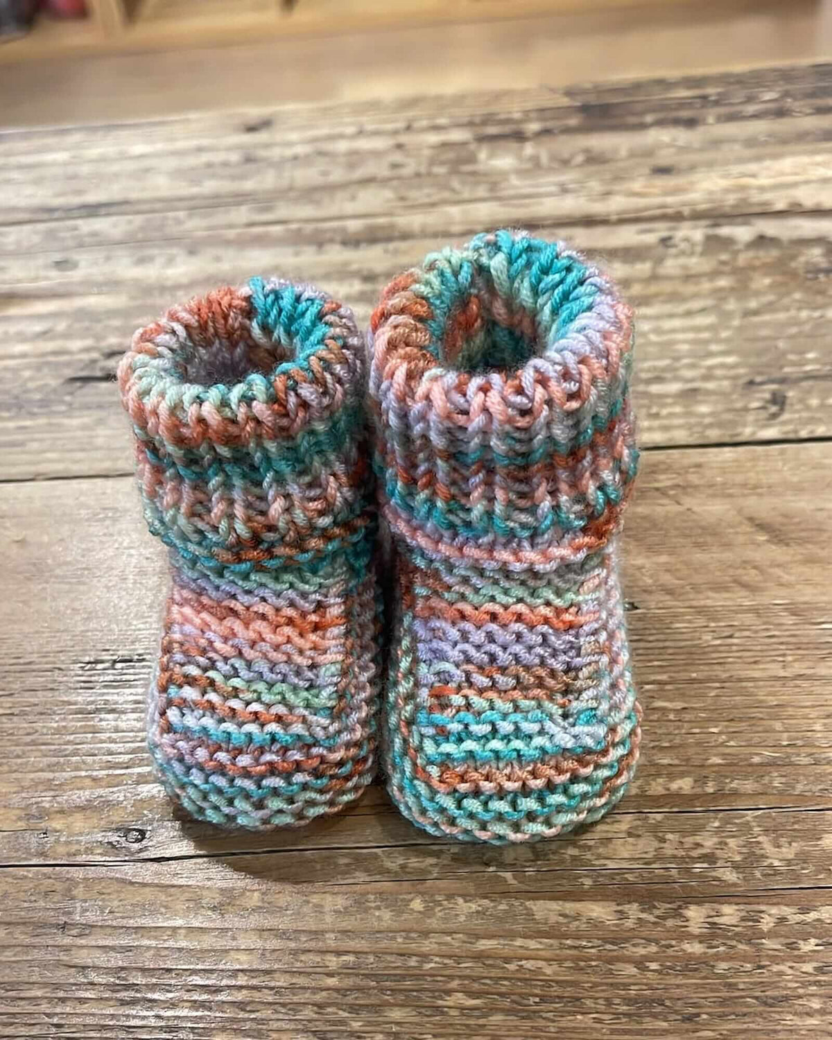 Hand-knit baby booties in a variegated yarn with stripes of teal, peach, pink, and white, displayed on a rustic wooden surface.