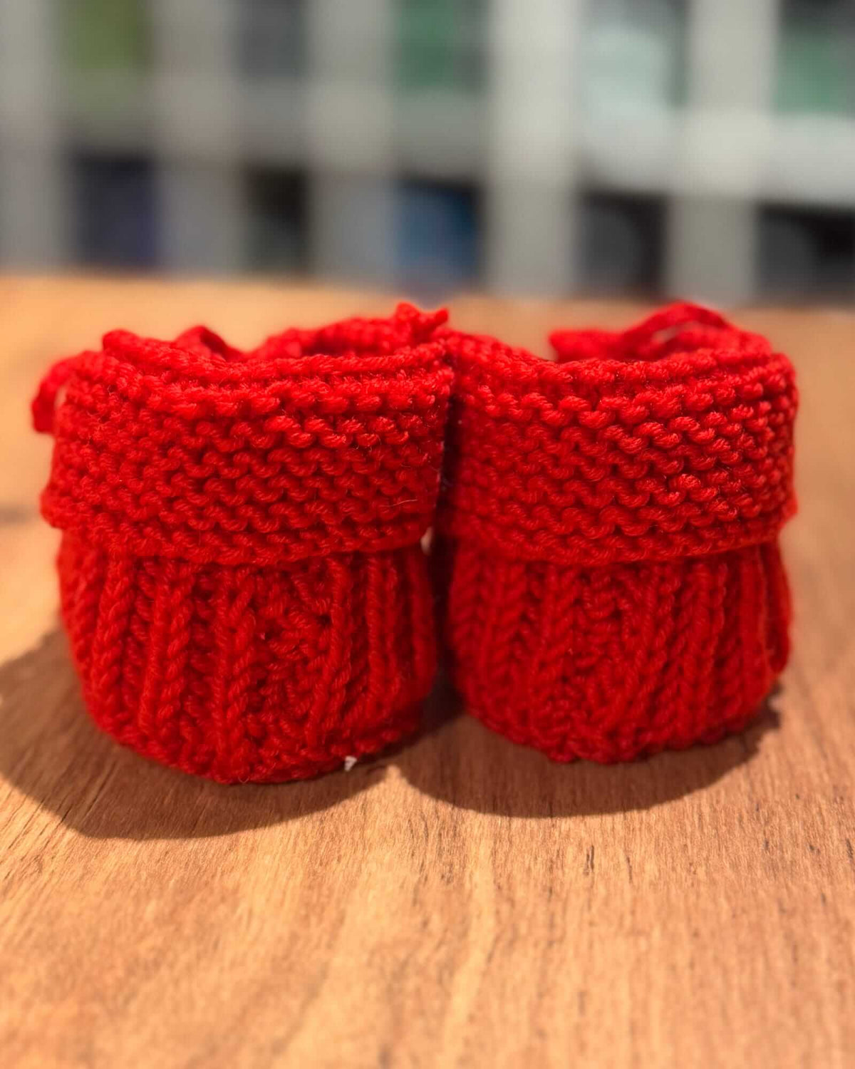 A pair of bright red hand-knit baby booties, shown from behind, resting on a wooden tabletop with a blurred indoor background.