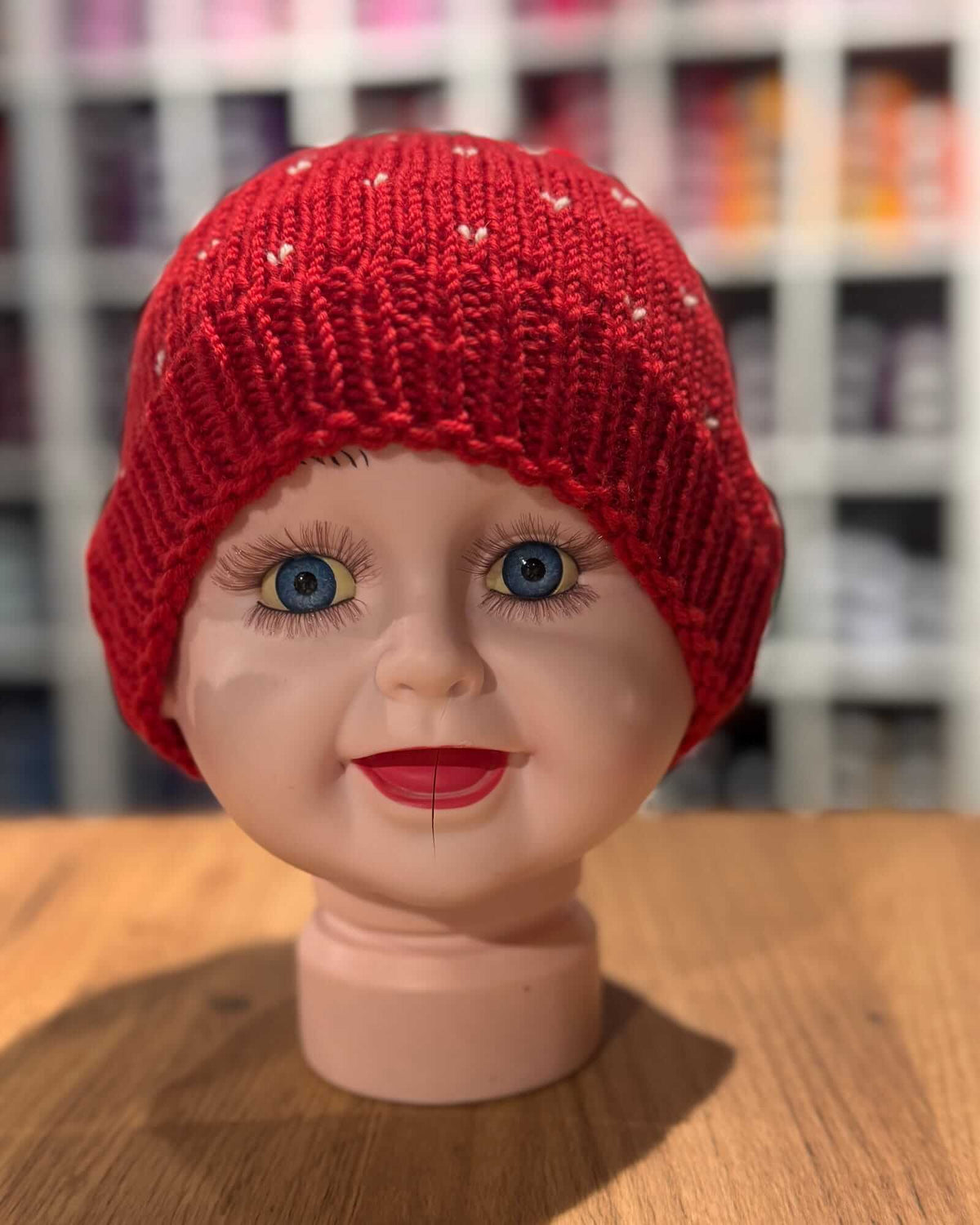 A close-up of a bright red knit beanie hat displayed on a doll head mannequin, sitting on a wooden surface with a blurred store background.