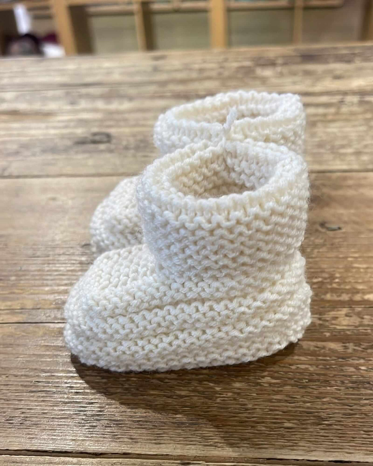 A pair of off-white hand-knit baby booties resting on a rustic wooden plank surface.