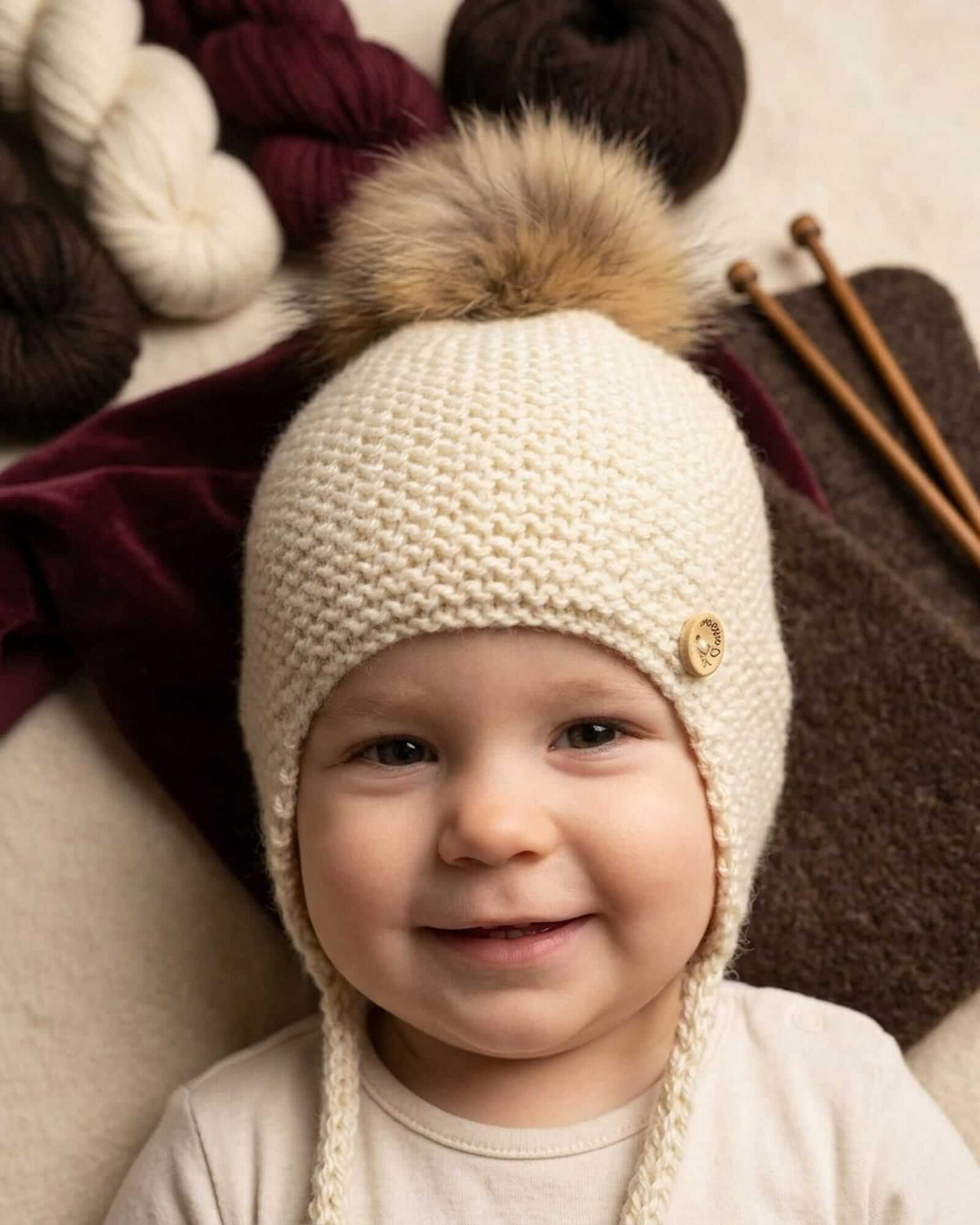 A baby wearing a cream-colored knit baby bonnet with a brown faux fur pom-pom on top, displayed on a doll's head. The bonnet features a small decorative button on the side and a chin strap, set against a background of cream, burgundy and chocolate coloured knitting stuff.