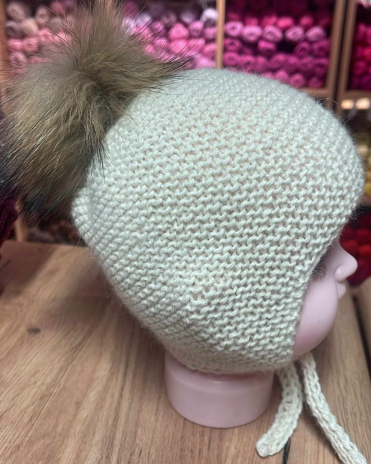 A light beige knit winter hat with ear flaps and a large brown faux-fur pom-pom, displayed on a pink mannequin head on a wooden surface.